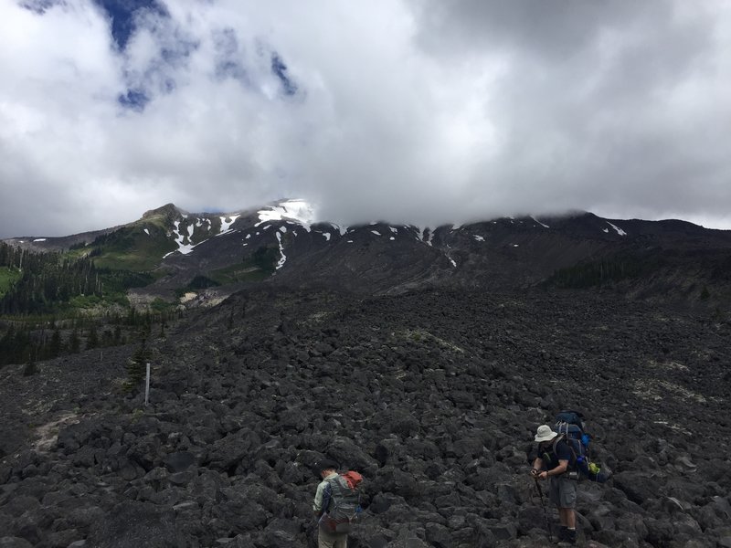 A couple of hikers on the "trail" with Mt St. Helens in the background obscured by clouds.