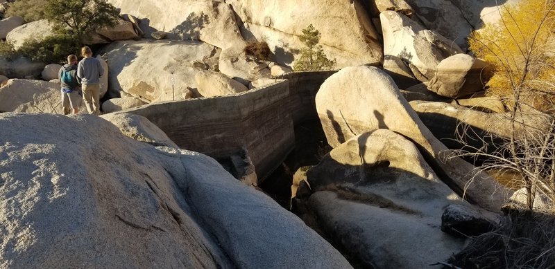 Westside or dam wall of Baker Dam picture center. Note water level even after a rain is low, Dec 2018. Trail continues though boulders screen left and behind view. People were hiking very close to the wall but rangers were around.