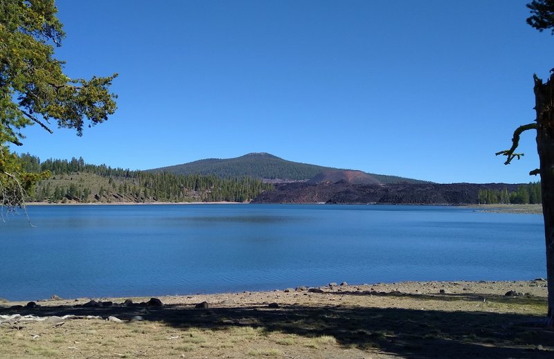 Prospect Peak, 8,338 ft., is the forested mountain in the distance. In front of Prospect Peak's right shoulder is Cinder Cone, and in front of Cinder Cone are the black boulders of Fantastic Lava Beds. Seen looking northwest across Snag Lake.