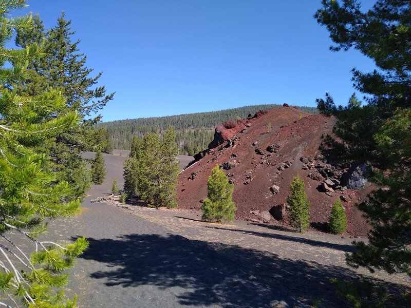 Fantastic Lava Beds coated with red, oxidized volcanic ash/cinders on the right.  In the distance behind Fantastic Lava Beds is the gently sloped, forested Prospect Peak, an ancient shield volcano.