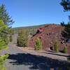 Fantastic Lava Beds coated with red, oxidized volcanic ash/cinders on the right.  In the distance behind Fantastic Lava Beds is the gently sloped, forested Prospect Peak, an ancient shield volcano.