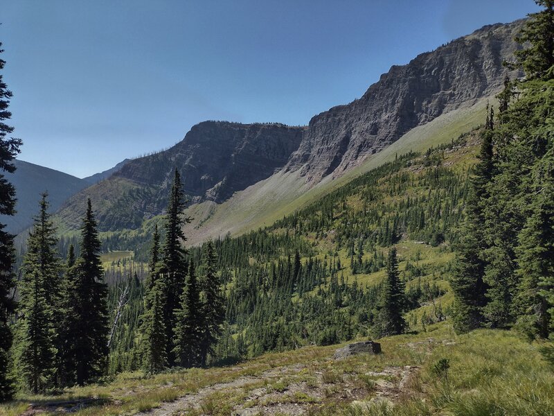 Rock walls of the Great Divide with meadows below, as the trail heads to a cute tarn in the distance on the left.