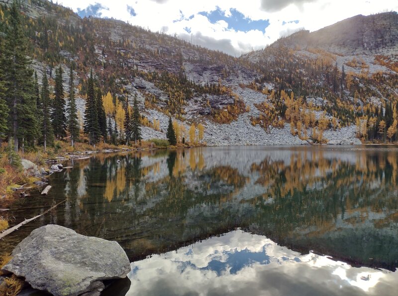 Rocky slopes, golden larch, firs and sky are perfectly reflected in the first lake during a calm moment.