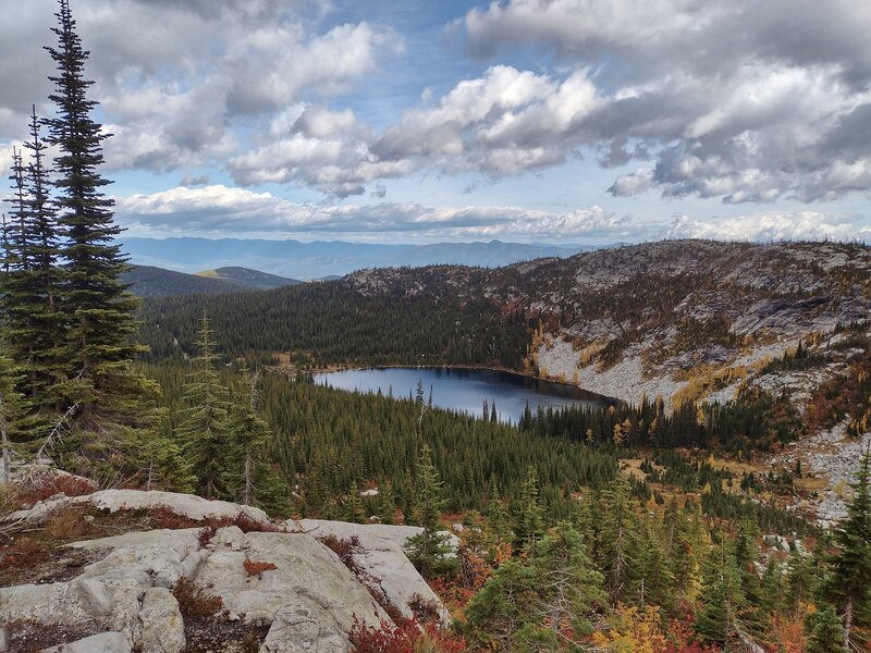 The lower/first Roman Nose Lake sparkles below in its boulder strewn bowl. In the distance (left) to the east, beyond the Kootenai River Valley, the Purcell Mountains meet the clouds and sky on the horizon.