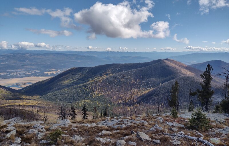 Farnham Ridge, front and center, with Farnham Peak, 6,907 ft., as its high point, radiates out from the ridge that leads to Fisher Peak. The Kootenai River (left) in its broad valley is in the distance below. Beyond are Purcell Mountains. All to the east.