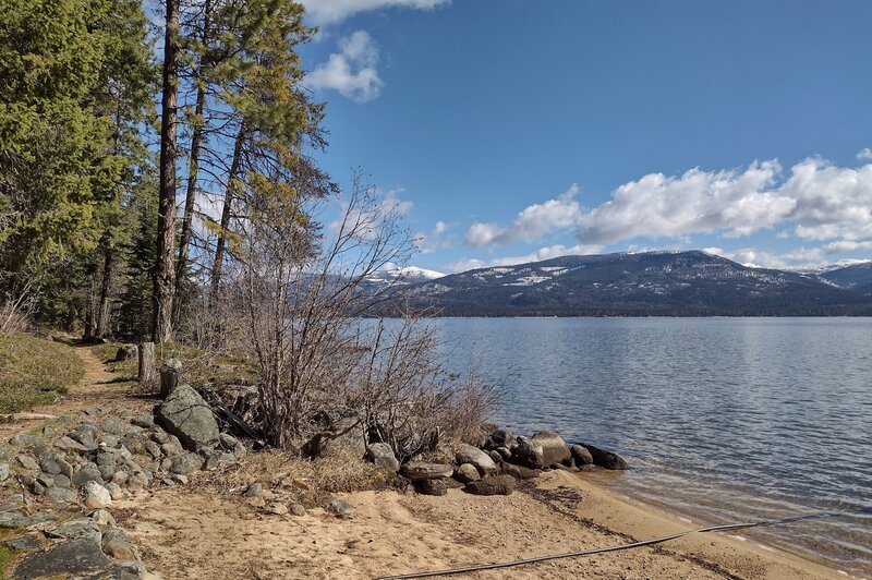 Cameis Prairie is the nearby hill across Priest Lake. Mount Roothaan (center) is the snowy peak in the distance. Seen looking northeast from one of the sandy beaches along Beach Trail.