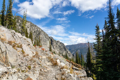 Navigating a rugged granodiorite shelf along the Chimney Lake Trail at 7,537 ft, the path skirts towering glaciated crags as the timber parts to reveal dizzying views into the Lostine River Valley.