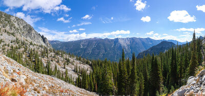 From a panoramic granodiorite shelf along the Chimney Lake Trail at 7,587 ft, a grand alpine vista sweeps across the glacially carved chasm of the Lostine River Valley toward the towering crests of the Hurricane Divide.