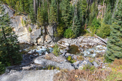 Looking down into the rugged bedrock gorge of the West Fork Wallowa River from the trail at 5,426 ft, crystal-clear rapids and deep pools churn through jointed granodiorite boulders approximately 70 ft below.